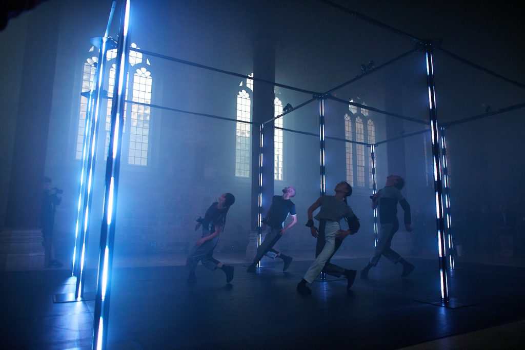 Four dancers in Strange Stranger. They are in a square formation, with the light towers of the installation gleaming.