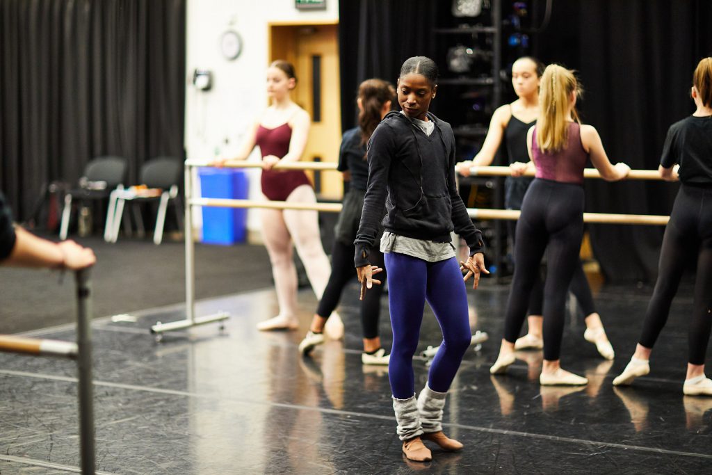 A Ballet Black dancer leads a balelt technique class. She is watchign the students at the barre, her fingers outstretched at her sides.
