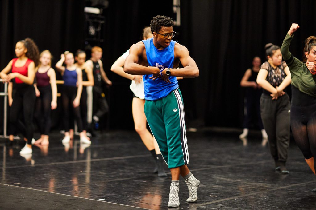 A Ballet Black dancer is leading a workshop with students. The back of his hands are meeting at his chest, with his elbows pointed out either side. One of his feet is crossed in front of the other.