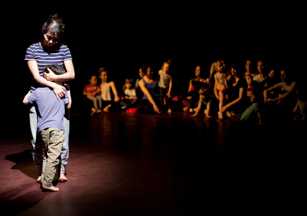 A performance of Touch by Four Hands. A dancer stands over a child, cradling the child's head in their handss. We can see the audience in the background.